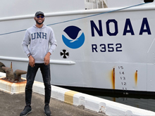 Elias Adediran stands on the pier beside the NOAA Ship Nancy Foster wearing a UNH hoodie.