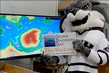 UNH mascot Gnarlz( in wildcat costume head and UNH hockey jersey) poses with ODD sign in front of a monitor showing a bathymetric projection of a sandbox.
